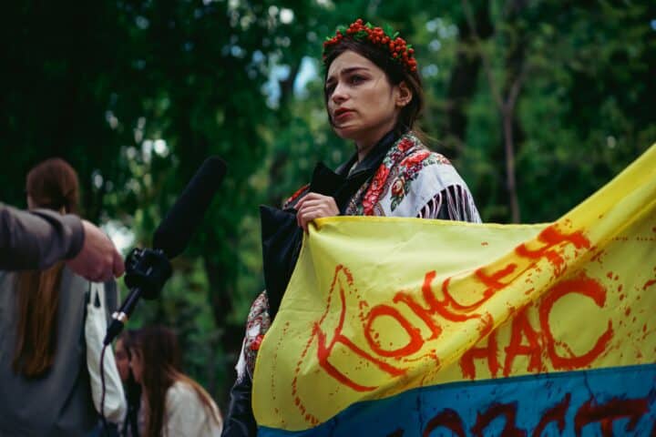 A woman holds a bloodstained flag of Ukraine in her hands, dressed in traditional Ukrainian clothing, protesting the war. Someone is holding a microphone close to her.